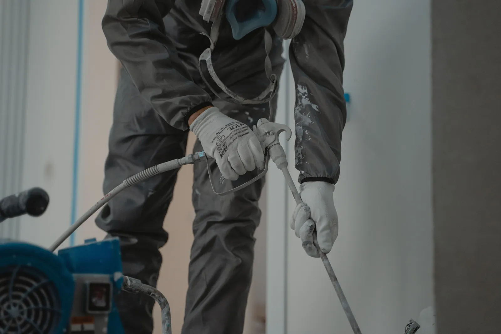 Close-up of gloved hands holding an airless spray gun and extension wand, with a blue paint sprayer in the foreground