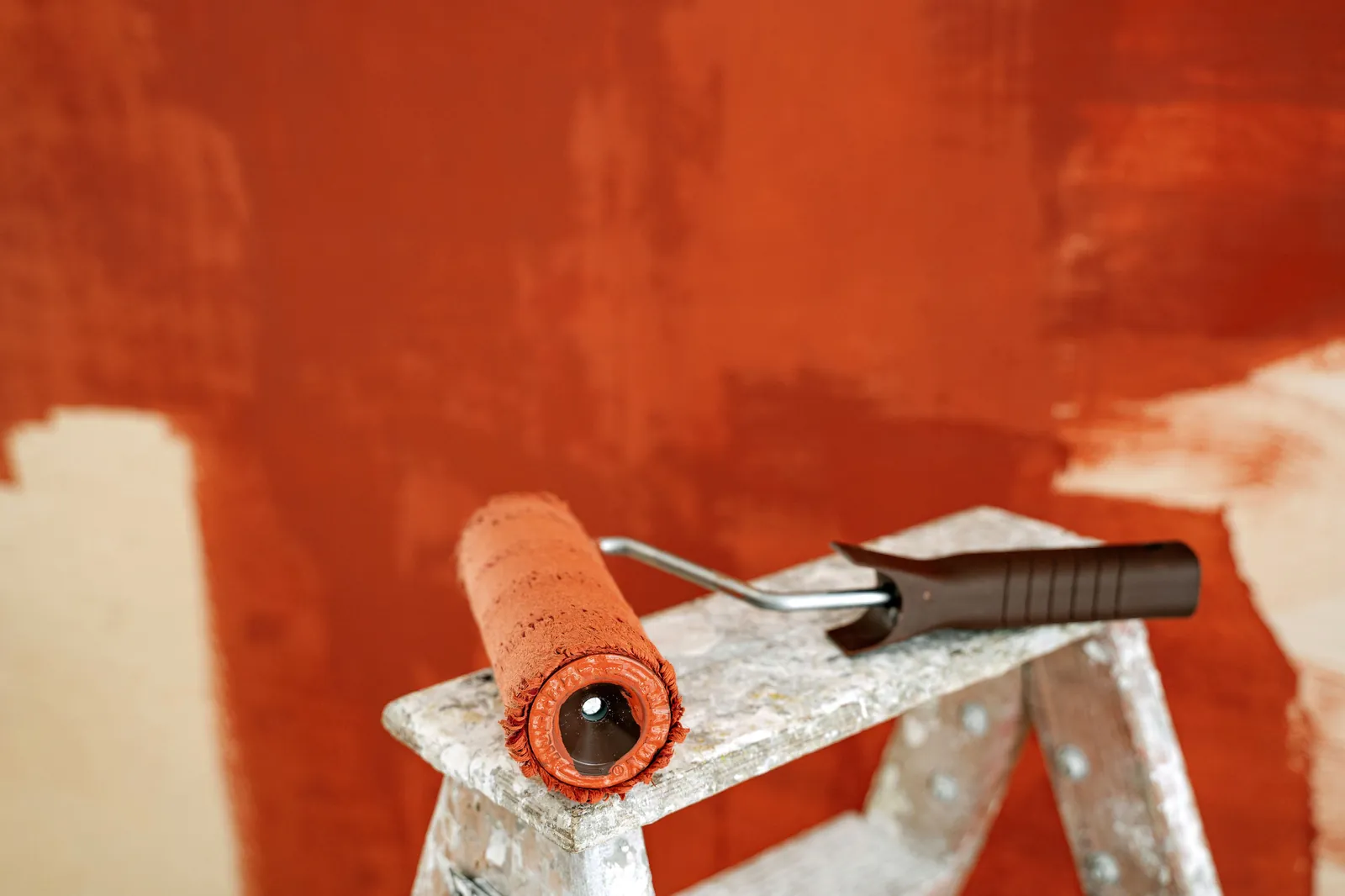 A paint roller on a stepladder Paint roller with an orange cover resting on a paint-spattered stepladder in front of a partly painted terracotta wall