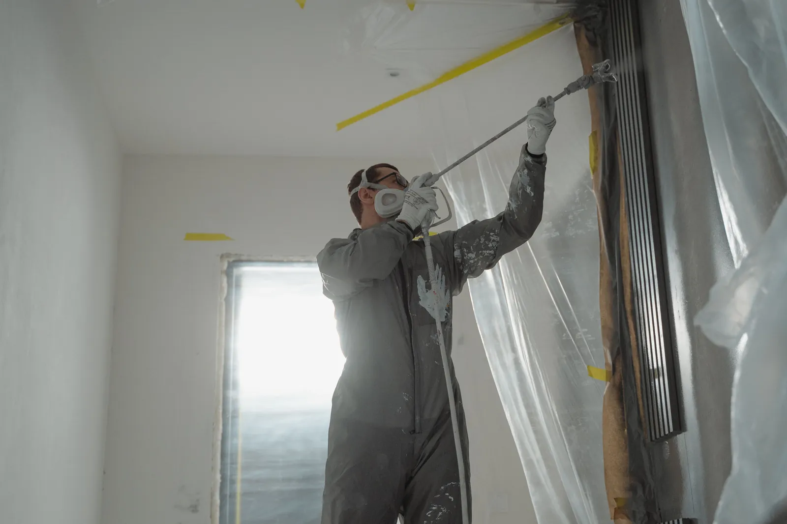 Painter in a respirator spraying the top of a wall with an extension wand, in a room covered in plastic sheeting and yellow tape